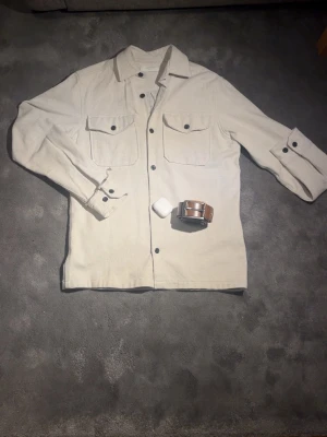 Beige overshirt från Jack & Jones - Stilren beige overshirt från Jack & Jones Premium med två bröstfickor och svarta knappar. Overshirten har klassisk krage och är tillverkad i ett kraftigare tyg som ger en snygg och avslappnad look. Perfekt att bära öppen över en t-shirt.✅
