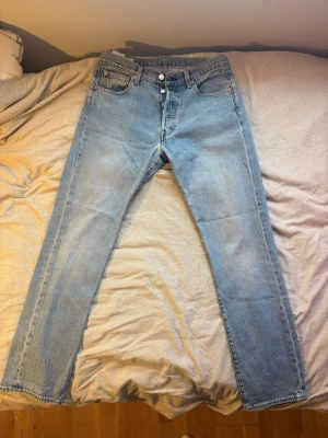 Levi's ljusblå 501 stl 30,32 - Säljer ett par klassiska ljusblå jeans från Levi's med raka ben och. Jeansen har fem fickor, knäppning med knappar och den ikoniska läderlappen bak i midjan. Tillverkade i slitstark denim, perfekta för en avslappnad och tidlös stil.