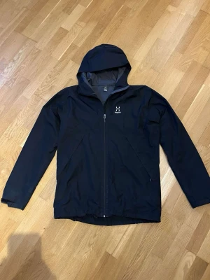 Svart vindjacka från Haglöfs med huva - Snygg svart vindjacka från Haglöfs med hel dragkedja framtill och praktisk huva. Jackan har två sidofickor med dragkedja och diskret Haglöfs-logga på bröstet. Tillverkad i ett lätt och slitstarkt material som passar perfekt för blåsiga dagar.