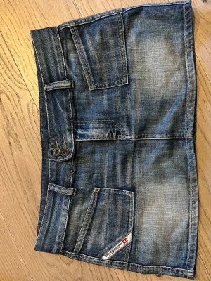 Blå jeanskjol från Diesel - Säljer en snygg blå jeanskjol från Diesel med slitna detaljer och stora fickor både fram och bak. Kjolen har bälteshällor och två knappar i midjan. Perfekt för dig som gillar en avslappnad och trendig stil.