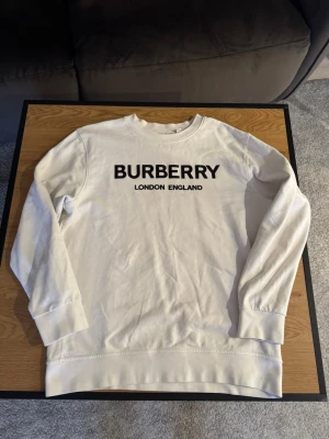 Vit sweatshirt från Burberry - Snygg vit sweatshirt från Burberry med svart tryck på bröstet där det står 'BURBERRY LONDON ENGLAND'. Tröjan har rund halsringning, långa ärmar och breda muddar vid ärmslut och nederkant. Perfekt för dig som gillar stilrena och exklusiva plagg.