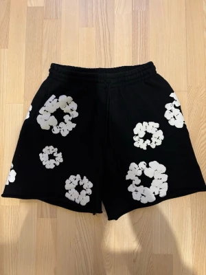 Svarta shorts från Denim Tears med blommor - Svarta shorts från Denim Tears med vita blommiga tryck över hela plagget. De har elastisk midja och avslappnad passform, perfekta för en chill och trendig stil. Materialet känns mjukt och bekvämt, troligen bomull.