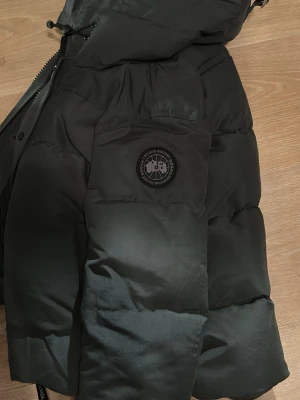 Svart dunjacka från Canada Goose 1:1 - Säljer en mörk grå dunjacka från Canada Goose med klassisk patch på ärmen. Jackan har huva, två sidofickor med dragkedja och ribbade muddar vid ärmsluten. Perfekt för kalla vinterdagar och riktigt varm tack vare dunfyllningen.