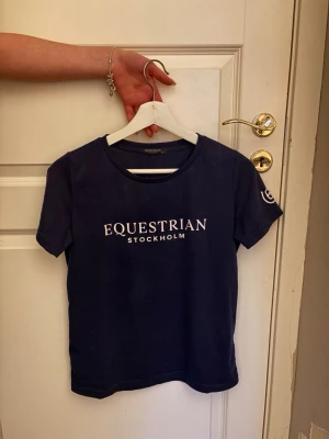 Mörkblå t-shirt Equestrian Stockholm - Mörkblå t-shirt från Equestrian Stockholm med vit logga tryckt på bröstet. Klassisk rund halsringning och korta ärmar. Perfekt för dig som gillar stilrena och sportiga plagg. Priset går att diskuteras och den är i storlek xs-s. 