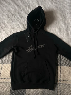 Svart Saint Laurent hoodie med tryck - Svart hoodie från Saint Laurent med coolt tryck på bröstet i grått och vitt. Klassisk känguruficka framtill och justerbar huva med snören. Tillverkad i mjukt material som känns skönt mot huden. Perfekt för en avslappnad streetwear-look.