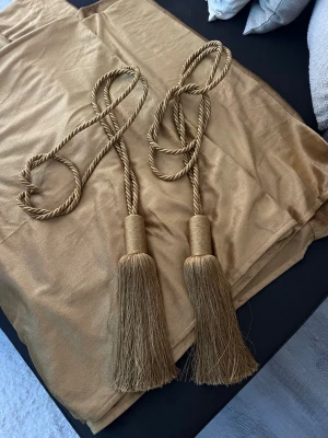 Guldbruna sammetsgardiner med omtag från Mio - Säljer ett par guldbruna sammetsgardiner tillsammans med två matchande gardinomtag med dekorativa tofsar. Gardinerna har en mjuk och lyxig känsla och tofsarna är i tvinnat snöre med långa fransar. Perfekt för att ge rummet en elegant touch. 2.50x1.40