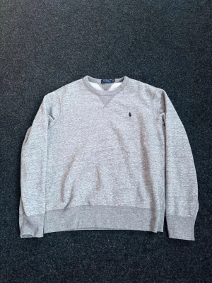 Grå sweatshirt från Polo Ralph Lauren - Klassisk grå sweatshirt från Polo Ralph Lauren med rund halsringning och diskret broderad logga på bröstet. Tröjan har ribbade muddar vid ärmslut och nederkant, perfekt för en avslappnad och stilren look.