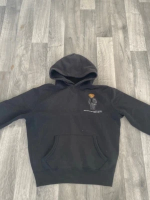 Svart hoodie Polo Bear Ralph Lauren - Svart hoodie från Ralph Lauren med Polo Bear-brodyr på bröstet. Klassisk känguruficka framtill och huva. Mjuk bomullskänsla och avslappnad passform, perfekt för en chill look.
