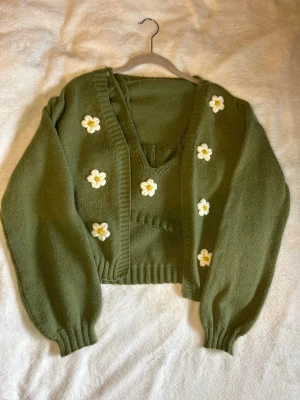 Olivgrönt stickat set med blommor - Sött stickat set i olivgrönt med en kofta och matchande crop top. Båda plaggen har virkade vita och gula blommor som dekoration. Koftan har ballongärmar och ribbade muddar, medan toppen har smala axelband och v-ringning. 