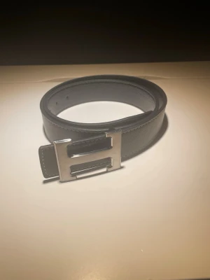 Svart skärp med silverfärgat H-spänne - Snyggt svart bälte med ett stort, silverfärgat H-format spänne. Bältet har en stilren och enkel design som passar till många olika outfits. Tillverkat i slätt skinn med diskreta sömmar längs kanterna.