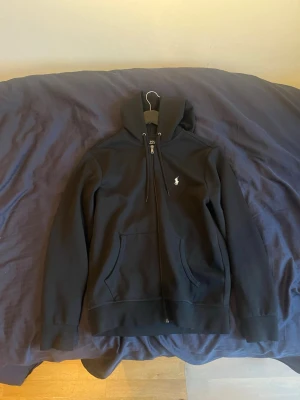 Svart zip hoodie från Polo Ralph Lauren - Säljer en svart hoodie med dragkedja från Polo Ralph Lauren. Klassisk design med huva, fickor framtill och den ikoniska vita logon broderad på bröstet. Perfekt för en chill och stilren look. Tillverkad i mjukt material för extra komfort.