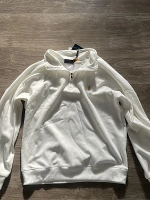 Vit quarter zip från Polo Ralph Lauren - Vit långärmad quarter zip-tröja från Polo Ralph Lauren med klassisk krage och dragkedja framtill. Broderad gul logga på bröstet. Perfekt för en clean och stilren look.