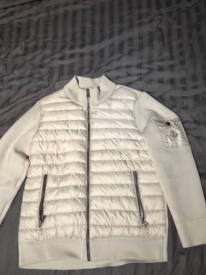 Vit cardiganjacka från Moncler - Snygg beige cardiganjacka från Moncler med quiltad framsida och stickade ärmar. Jackan har två dragkedjefickor framtill och en Moncler-logga på ärmen. Perfekt för lager-på-lager och stilrena outfits. Har använt den ett fåtal gånger och har några icke märkbara fläckar som kan tas bort.