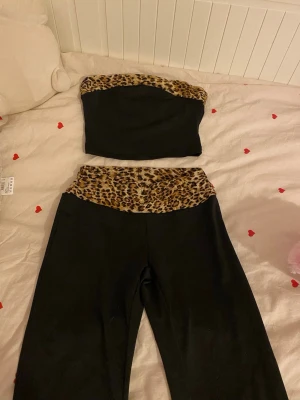 Svart set med leoparddetaljer - Tvådelat set med svart bandeau-topp och utsvängda byxor, båda med leopardmönstrade kanter. Setet har en trendig och ungdomlig look med djurmönster som kontrast. Perfekt för dig som gillar att sticka ut med din stil.