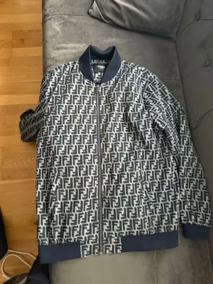 Fendi bomberjacka med FF-mönster - Snygg bomberjacka från Fendi med klassiskt FF-logomönster i svart och vitt. Jackan har ribbade muddar, krage och nederkant i mörkblått samt dragkedja framtill. Perfekt för dig som gillar streetwear och vill sticka ut med en ikonisk design.
