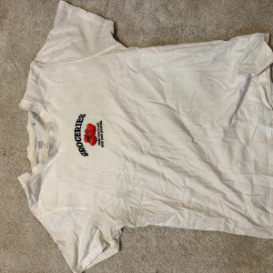 Vit t-shirt med tryck från 157 - Vit t-shirt från 157 med trycket 'GROCERIES' och två röda tomater på bröstet. Klassisk rund hals och korta ärmar. Tillverkad i 100% bomull, perfekt för en avslappnad och enkel stil.