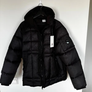 Svart pufferjacka från C.P. Company - Säljer en svart pufferjacka från C.P. Company med huva och dragkedja framtill. Jackan har en patch med logga på ärmen och två sidofickor. Riktigt fluffig, perfekt för kalla dagar. Materialet är glansigt och quiltat för extra värme. Storlek XXL asiatisk, som L europeisk.