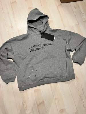 Grå hoodie från Enfants Riches Déprimés - Säljer en grå hoodie från Enfants Riches Déprimés med svart text på bröstet. Hoodien har huva, magficka och en oversized passform. Distressed detaljer och råa kanter ger en edgy vibe. Tillverkad i mjuk bomull för maximal komfort. 0bs lnte 0rglnal‼️, men gjord i mycket bra kvalitet 