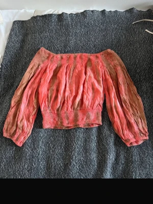 Corallfärgad offshoulder blus med puffärmar - Trendig corall färgad offshoulder blus med bred resår i midjan och långa puffärmar. Blusen  är perfekt för dig som gillar en avslappnad men ändå snygg stil. Färgen går i olika corall rosa orange  och beige toner.