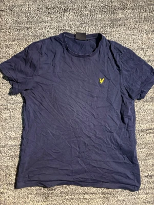 Mörkblå t-shirt från Lyle & Scott - Snygg mörkblå t-shirt från Lyle & Scott med klassisk gul logga på bröstet. T-shirten är kortärmad och har en enkel, stilren design i mjuk bomull. Perfekt till jeans eller shorts för en clean look.