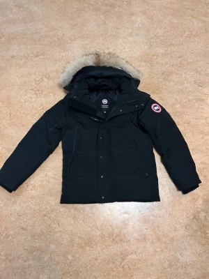 Svart dunjacka från Canada Goose - Säljer en svart dunjacka från Canada Goose med avtagbar huva och pälskant. Jackan har klassisk patch-logga på ärmen, dragkedja och knappar framtill samt två fickor med dragkedja. Köptes december 2025. Använd men inget slitage