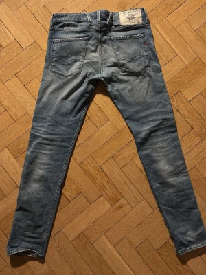 Blå slim fit jeans från replay. - Säljer ett par blå slim fit jeans från Replay Jeans i modellen anbass, med klassisk femficksmodell och snygga slitningar framtill. Jeansen har en ljus tvätt och detaljerade sömmar på bakfickorna. Perfekta för en avslappnad och trendig look.
