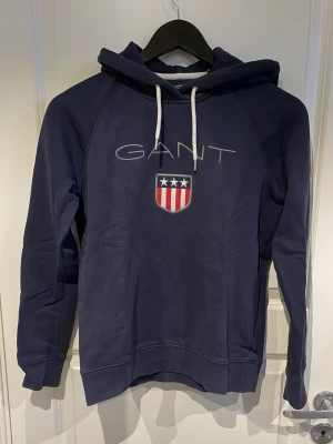 Mörkblå hoodie från GANT - Mörkblå hoodie från GANT med vit dragsko i huvan och stort tryck på bröstet med GANT-logga och amerikanskinspirerat sköldmärke i rött, vitt och blått. Liten i storleken och mjukt material, perfekt för en avslappnad stil.