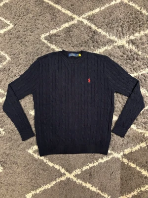 Mörkblå Polo Ralph Lauren tröja - Mörkblå kabelstickad tröja från Polo Ralph Lauren med rund halsringning och röd broderad logga på bröstet. Tröjan har långa ärmar och ribbade muddar vid ärmslut och nederkant. Jag är 180cm och väger 180kg och den passar mig perfekt.