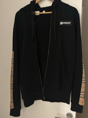 Svart Burberry hoodie med rutiga detaljer - Snygg svart hoodie från Burberry med dragkedja och klassiska beige-rutiga detaljer längs ärmarna. Märkeslogga på bröstet och huva med snören. Perfekt för dig som vill ha en stilren men ändå unik look. Tillverkad i mjukt bomullsmaterial.. storlek xs men passar mig som är medium 188 cm använd fåtal gånger kvitto finns, finns att hämta i Göteborg 