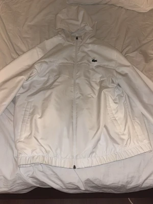 Vit vindjacka från Lacoste med huva - Snygg vit vindjacka från Lacoste med dragkedja framtill, två sidofickor och elastisk nederkant. Jackan har en huva och den klassiska Lacoste-loggan på bröstet. Perfekt för blåsiga dagar och enkel att matcha med olika outfits.