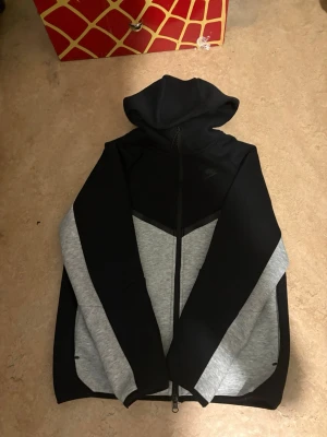 Nike svart och grått loungewear-set - Nike loungewear-set med hoodie och byxor i svart och grått. Hoodien har dragkedja och huva, medan byxorna har dragsko i midjan och fickor. Båda plaggen har snygga färgblock och Nike-logga. Perfekt för chill dagar hemma.