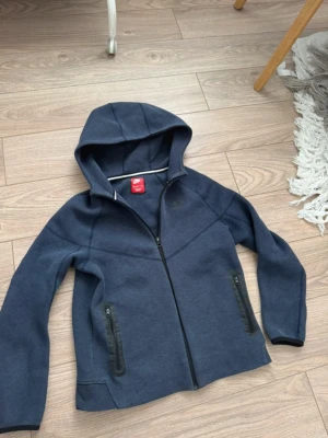 Mörkblå hoodie från Nike - Snygg mörkblå hoodie från Nike med dragkedja framtill och två fickor med svarta dragkedjor. Klassisk Nike-logga på bröstet och skön huva. Tillverkad i mjukt sweatshirtmaterial som passar perfekt till en avslappnad stil. Kan finnas tecken på användning men inga hål osv. Hör gärna av dig om du är osäker eller bara har någon fråga. Levereras alltid väl inpackat samt snabbt! Storlek 158-170 men sitter ungefär som en xs. Mvh. Sökord: nike, niketech,hoodie,vår,sommar,snygg.