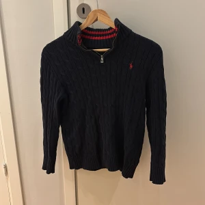 Mörkblå stickad tröja Polo Ralph Lauren - Mörkblå kabelstickad tröja från Polo Ralph Lauren med halv dragkedja och röd broderad logga på bröstet. Tröjan har ribbade muddar, snygga röda detaljer på insidan av kragen. Perfekt för kyliga dagar. Skick 9/10 nästan helt ny