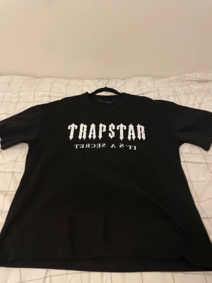 Svart Trapstar t-shirt Paris print - Svart t-shirt från Trapstar med vit text 'TRAPSTAR IT'S A SECRET' på framsidan och 'PARIS' i blått, vitt och rött på ryggen. Klassisk passform och rund hals. Perfekt för dig som gillar streetwear och vill sticka ut. Priset kan diskuteras 