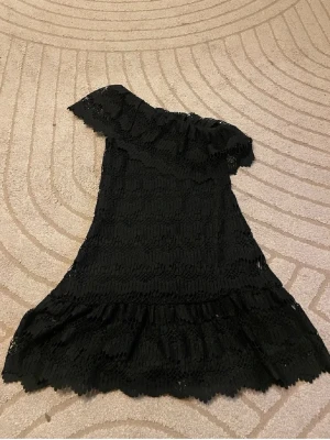 Svart spetsklänning med oneshoulder - Säljer en svart kortklänning med spets och volangdetaljer. Klänningen har oneshoulder-design och är tillverkad i spetsmaterial med vågigt mönster. Perfekt för dig som vill ha en unik och trendig look. Obs vita klänningen lånade bilder bara att se hur det ser ut på❤️