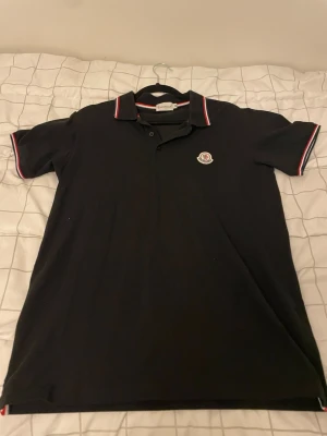 Svart Moncler pikétröja med logga - Klassisk svart pikétröja från Moncler med vit, röd och marinblå kant på krage och ärmslut. Tröjan har Monclers ikoniska logga broderad på bröstet och är tillverkad i mjukt bomullsmaterial. Perfekt för en stilren och sportig look. Priset kan diskuteras 