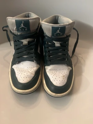 Nike Air Jordan 1 High vit/mörkgrön - Säljer ett par Nike Air Jordan 1 High sneakers i vit och mörkgrön färg. Klassisk siluett med högt skaft, perforerad tåbox och ikoniska Swoosh-loggan på sidan. Skorna har gröna snören och detaljer samt vit plös med Jumpman-logga. Tillverkade i läder.