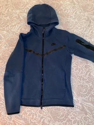 Blå Nike tech kofta - Snygg blå kofta från Nike med dragkedja framtill och huva. Tröjan har svarta detaljer över bröstet och på ena ärmen, samt diskret Nike-logga på bröstet. Perfekt för en sportig och avslappnad stil.