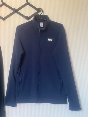Marinblå fleecejacka från Helly Hansen - Mysig marinblå fleecejacka från Helly Hansen med dragkedja hela vägen och två fickor med dragkedja. Jackan har hög krage och HH-logga på bröstet. Perfekt för lager-på-lager och svalare dagar.