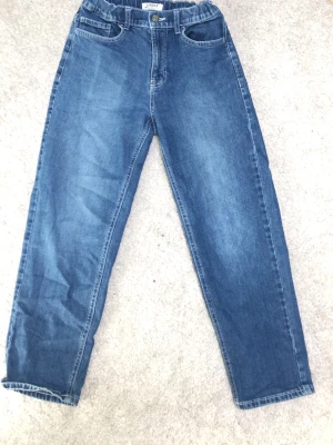 Blå jeans från Lindex, rak modell - Säljer ett par klassiska blå jeans från Lindex i rak modell. Jeansen har resår i midjan, knapp och dragkedja framtill samt två fickor fram. Perfekta för en avslappnad och stilren look.