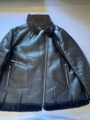 Svart skinnjacka med fuskpäls från Boohoo - Snygg svart skinnjacka från Boohoo med mjuk fuskpäls på insidan och kragen. Jackan har dragkedjor, två fickor framtill och detaljer i silver. Perfekt för dig som vill ha en stilren och trendig look. 