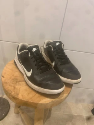 Svarta Nike sneakers med vit sula - Säljer ett par svarta sneakers från Nike med vit sula och vit swoosh-logga på sidan. Skorna har snörning och en sportig design, perfekta för inomhussport eller vardagsbruk. Ovandelen är i syntetmaterial och textil, med vadderad häl för extra komfort.
