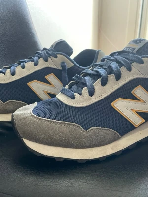 New Balance 515 blå sneakers - Snygga New Balance 515 sneakers i marinblått mesh med grå mockadetaljer och vita inslag. Klassisk N-logga i vitt och gult på sidan. Skorna har blå snören och en bekväm, vadderad sula. Perfekta för dig som gillar retrostil och streetwear.