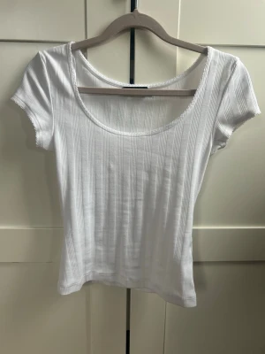 Snygg vit topp med rundad hals och snygga detaljer  - Säljer en vit topp med rund, djup urringning och korta ärmar. Toppen har en tight passform och söta detaljer samt är tillverkad i mjukt bomullsmaterial. Perfekt att styla med jeans eller kjol för en enkel och fräsch look. 💕 säljer då den tyvärr bara kommit till användning en gång. Pris kan diskuteras!