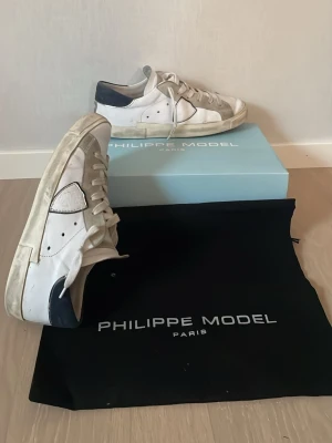 Philippe Model vita sneakers med blå detaljer - Snygga vita sneakers från Philippe Model med marinblå detaljer på hälen och sidan. Skorna har klassisk låg profil, vita skosnören och en robust sula. Materialet är en mix av läder och mocka för en cool och trendig look.