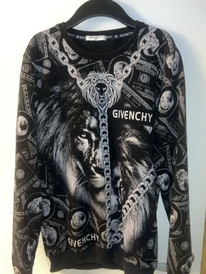 Givenchy svart sweatshirt med lejon - Svart sweatshirt från Givenchy med stort lejonmotiv, kedjedetaljer och dollartryck över hela tröjan. Tydlig Givenchy-logga både fram och bak. Tröjan har rund hals och ribbade muddar. Perfekt för dig som gillar statement-plagg och streetwear.