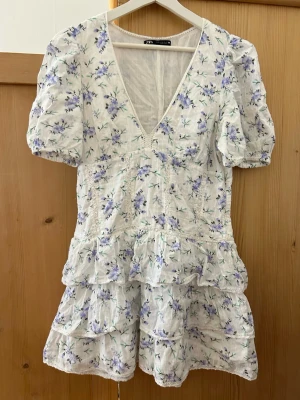 Zara volangklänning blomrig - Säljer denna supersöta och rare Zara-klänning som blivit väldigt trendig och svår att få tag på 💙🤍 Klänningen har puffärmar, djup v-ringning och flera volanglager som ger den perfekta feminina/coquette looken. Vit med ljusblå blommor och jättefin passform ✨  Storlek XL men passar bättre som L då den är mindre i modellen. Jag är storlek M/L. Använd som min studentklänning och endast använd få gånger, därför i superfint skick utan defekter 🌸
