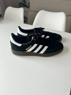 Adidas Spezial svarta sneakers - Svarta Adidas Spezial sneakers med klassiska vita ränder på sidorna och guldfärgad text. Skorna har svart mocka, vita detaljer vid hälen och bruna gummisulor. Snygg retrostil med låg profil och snörning framtill.