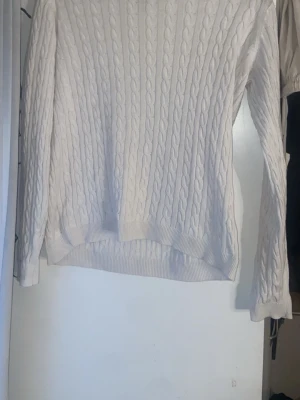 Vit kabelstickad tröja - Säljer en klassisk vit kabelstickad tröja med rund hals och långa ärmar. Tröjan har ribbade muddar vid ärmslut och nederkant, vilket ger en snygg och tidlös look. Perfekt att matcha med jeans eller kjol för en stilren outfit.