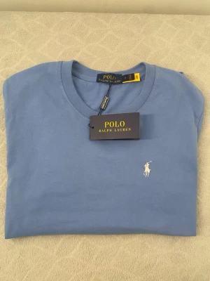 Blå t-shirt från Polo Ralph Lauren - Snygg blå t-shirt från Polo Ralph Lauren med klassisk rund halsringning och liten vit broderad logga på bröstet. Tillverkad i mjuk bomull som känns skön mot huden. Perfekt för en stilren och avslappnad look.